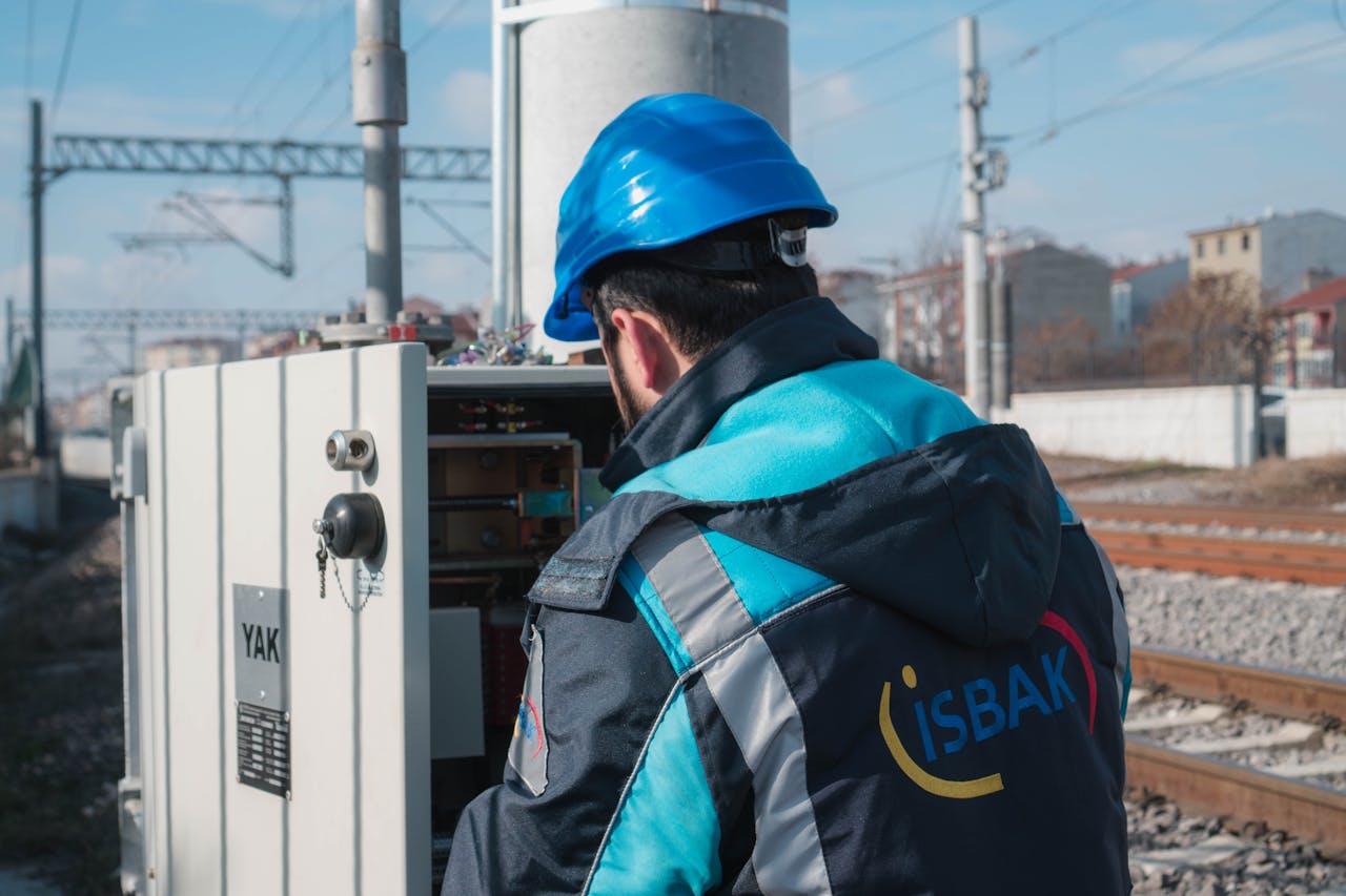 An engineer in a blue hard hat examines a fuse box on a railway track outdoors.